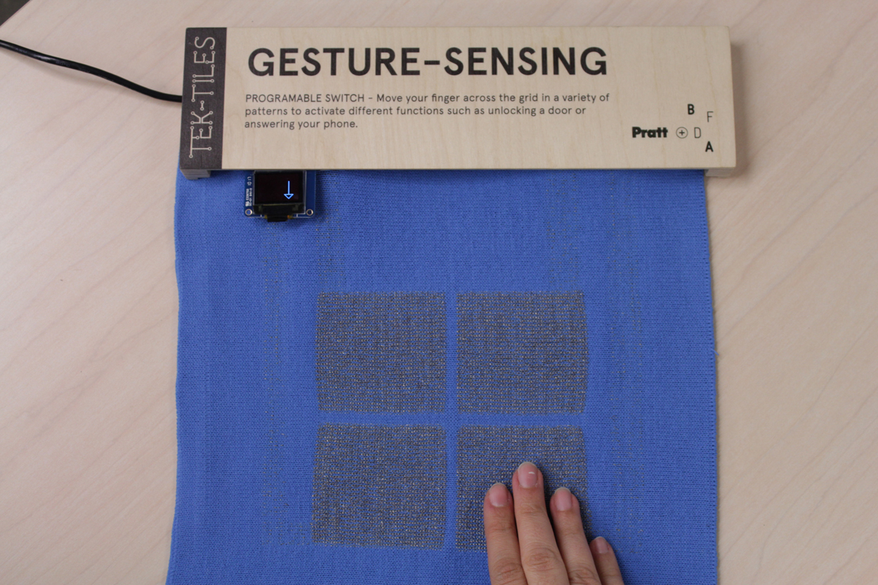 Photo of the knitted fabric on the exhibition hanger. There is a hand on the bottom right of the fabric, and on the top right there is a tiny screen with an arrow pointing down. The exhibition hanger is made of wood and has the following text: Tel-Tiles Gesture-Sensing Programmable Swicht – Move your finger across the gird in a variety of patterns to activate different functions such as unlocking a door or answering your phone. Pratt BFDA.