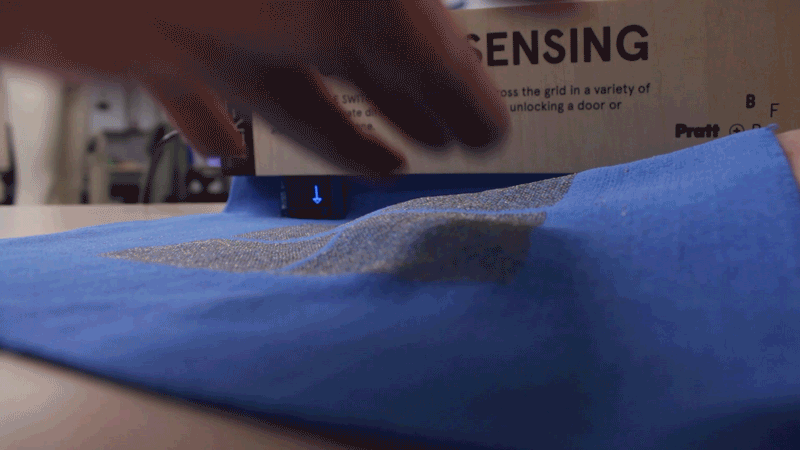Animated gif on loop showing a hand touch a fabric up and down. On the back there is a small screen showing an arrow pointing to the respective direction the fabric was being touched.