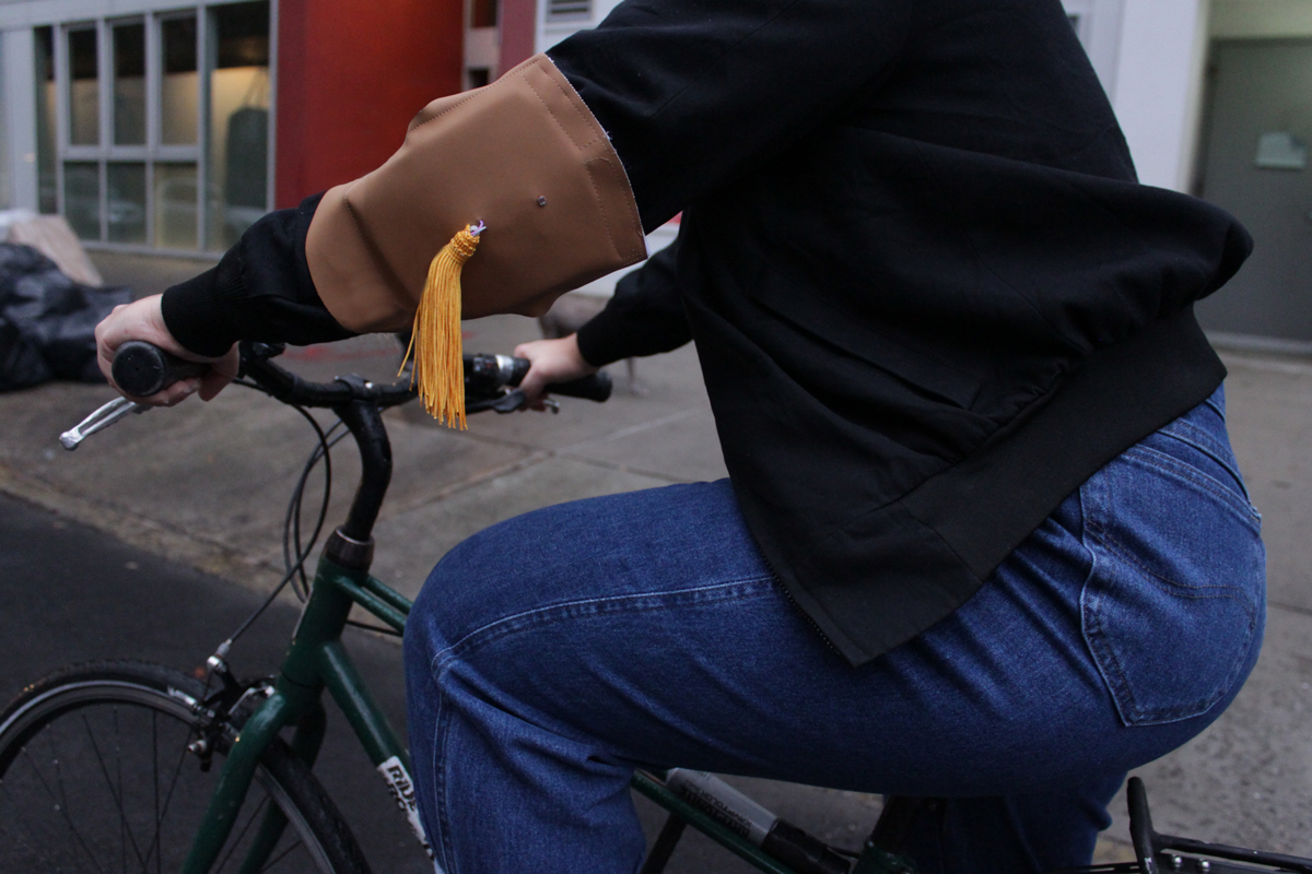 Photo of a women sitting on a bike. She is wearing the wearable device on her left shoulder. It is made out of beige faux leather and it has a yellow fringe falling from the middle of it.
