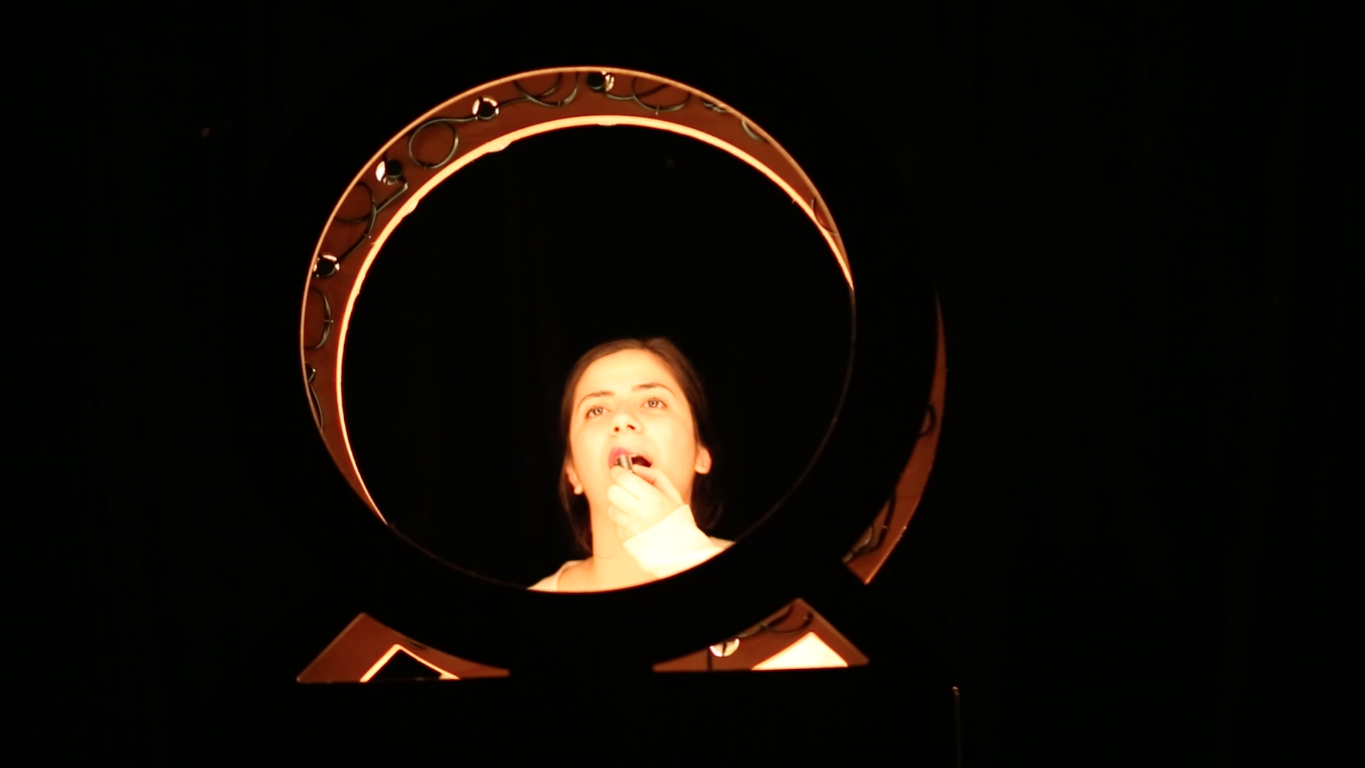 Photo of the perfomance. The background is black, the artist is behind a ring light that is illuminating her face and she is putting lipstick on.