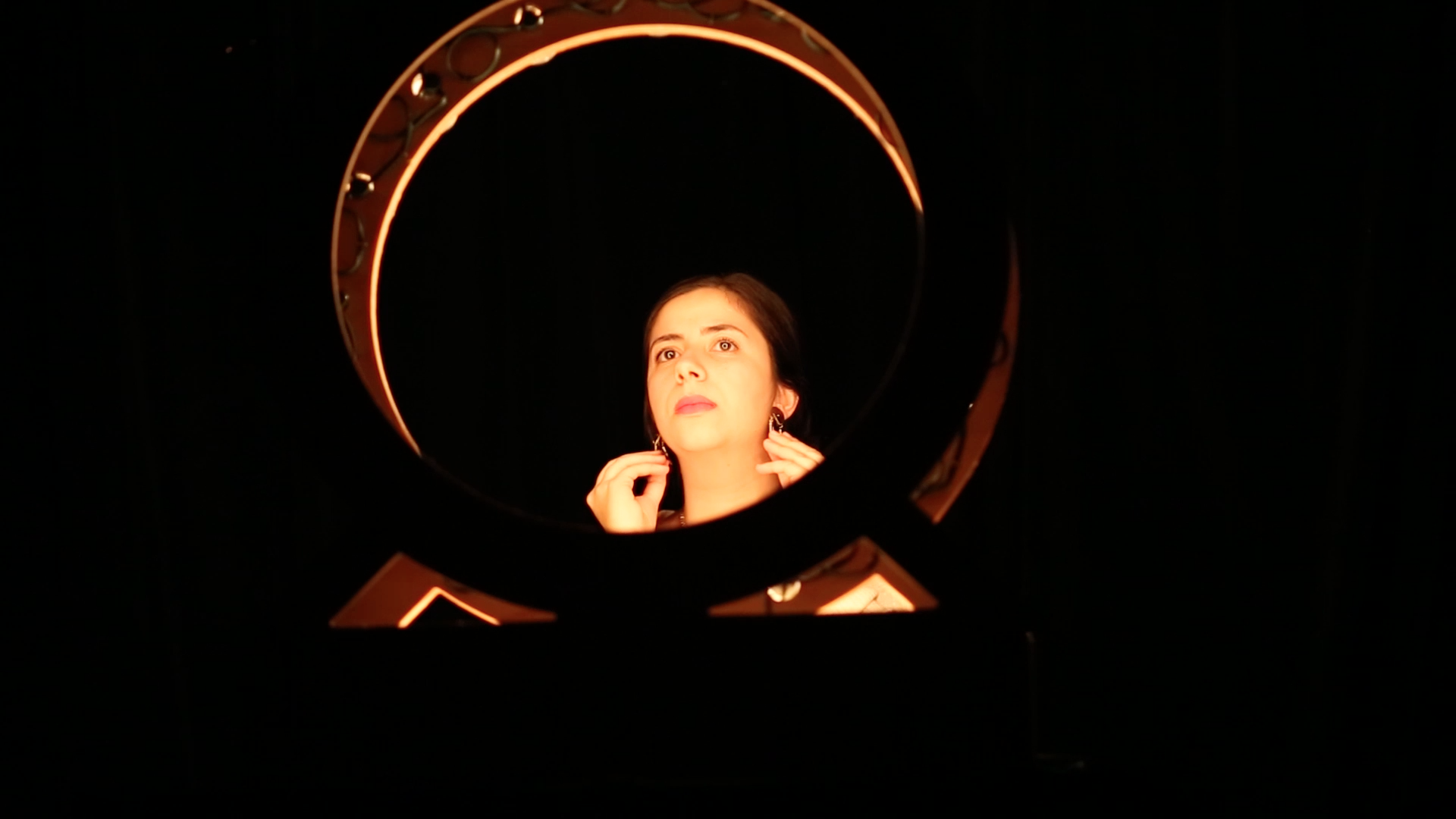 Photo of the performance. The artist is behind a ring light and she is putting earrings on.
