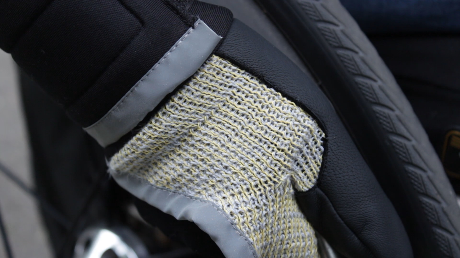 Photo with a detailed view of the glove. The hand wearing it is holding on the wheel of a wheelchair. The glove is knitted with yellow kevlar and grey cotton. the seams have a grey reflective fabric, and on the thumb there is a black leather reinforcement sewn on top of the knit.