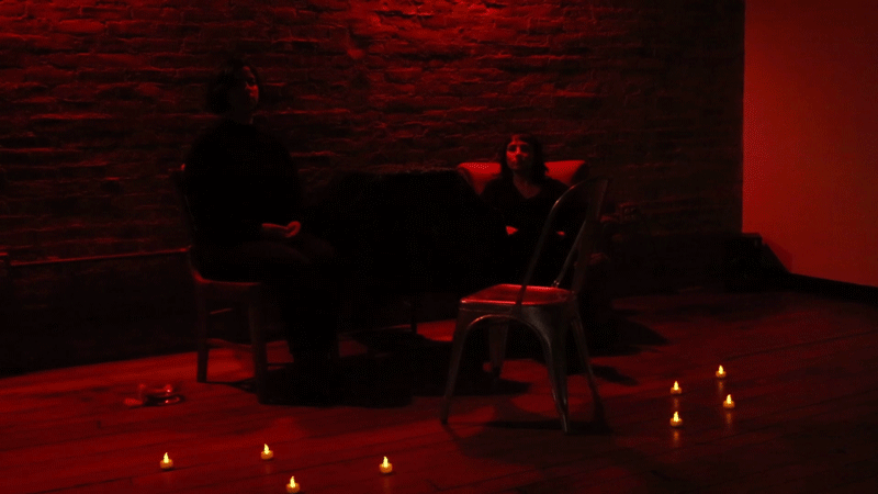 Photo of the installation’s setup. The room is illuminated by a red light, there are small candles spread on the floor. The two artists are sitting in front of an empty chair, and between them there is a table covered with a cloth.