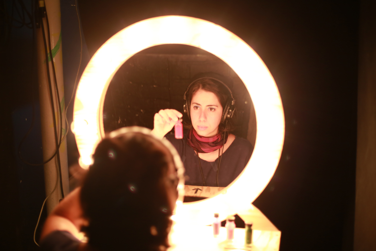 Photo of the artist activating the installation. Her image is reflected on the round mirror that has a ring light around it and the artists is wearing headphones and holding a tiny glass container with a pink liquid inside it.