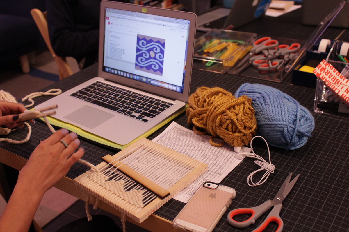 Photo of an attendee weaving a pattern. The pattern was programmed and is visible on the screen of the computer on the left of the picture. On the table you can also see a paper with the original drawn pattern and blue and yellow yarn balls.