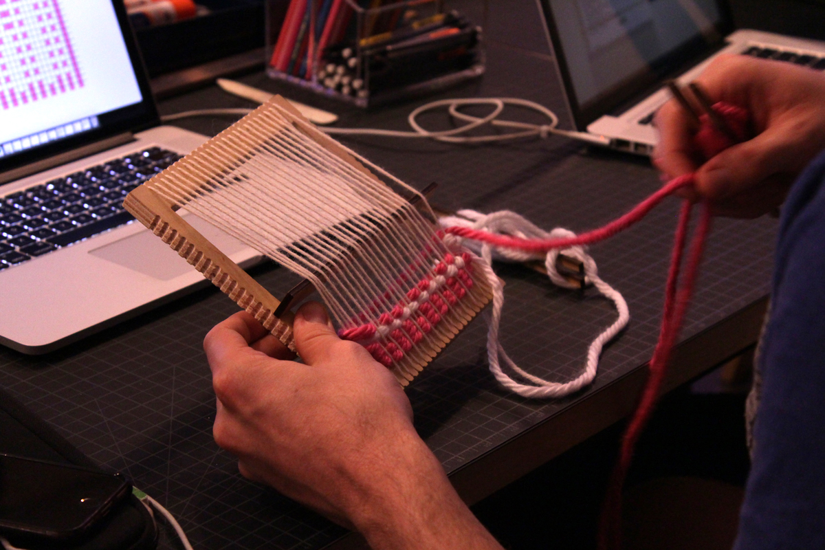 Photo of an attendee weaving a pattern. On the top left you can see a glimpse of the computer’s screen, with the coded pattern.
