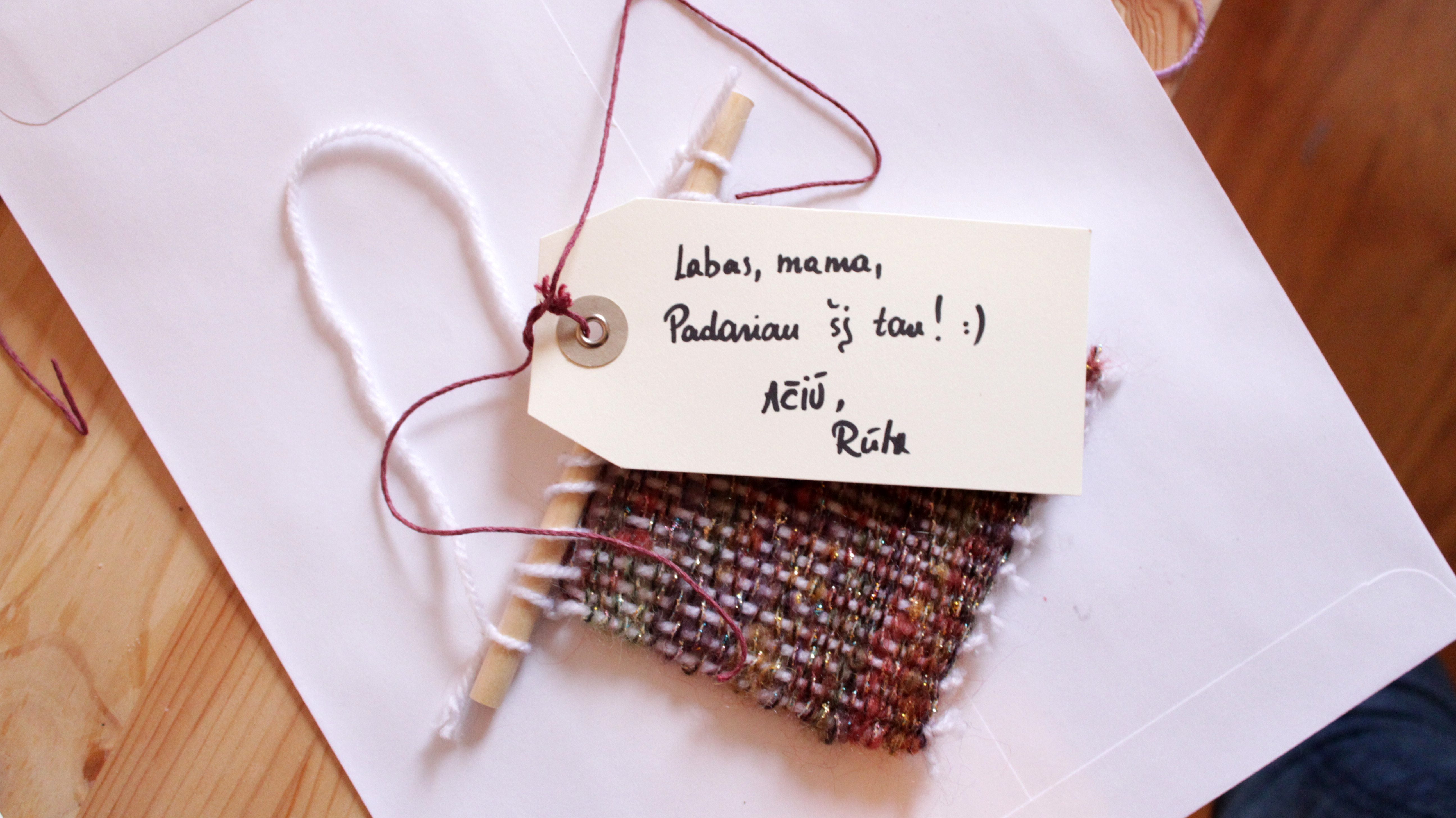 Photo of a weaved piece ready to be mailed. There is a tag on the top of it with a handwritten message in lithuanian and bellow it, a white envelope.