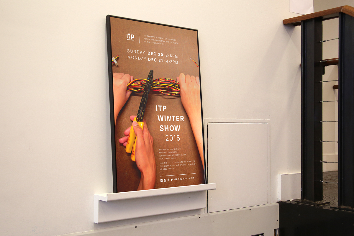 Photo of a framed poster for ITP Winter Show of 2015. The background of the poster is a photo in which there are two hands holding wires together, and a third one with a plier trying to cut them.
