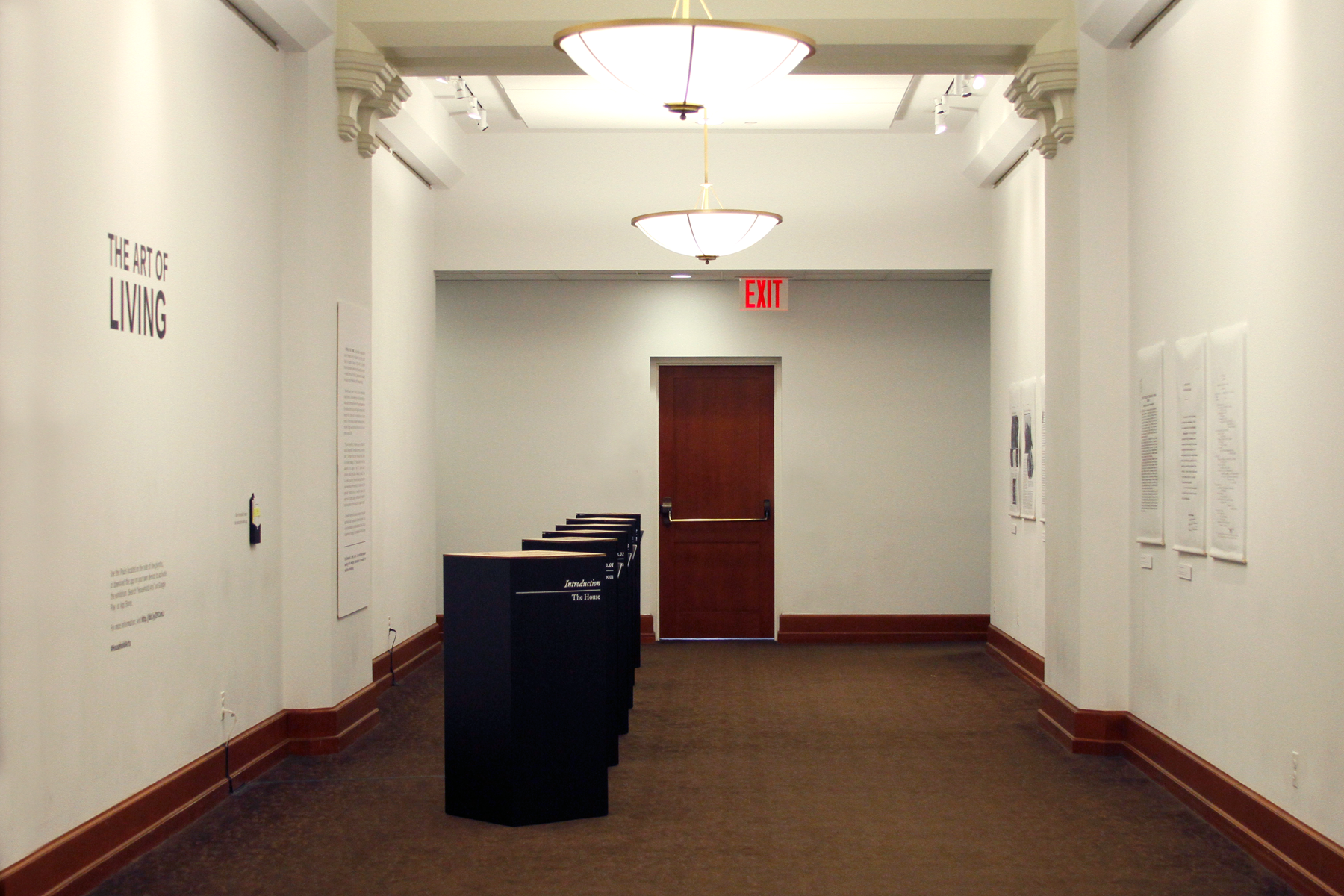 Photo of a hallway with the exhibition. On the left wall, the title and text about the exhibition. On the center-left of the hall there are six plinths lined up until the end of the walls. And on the right wall there are posters.